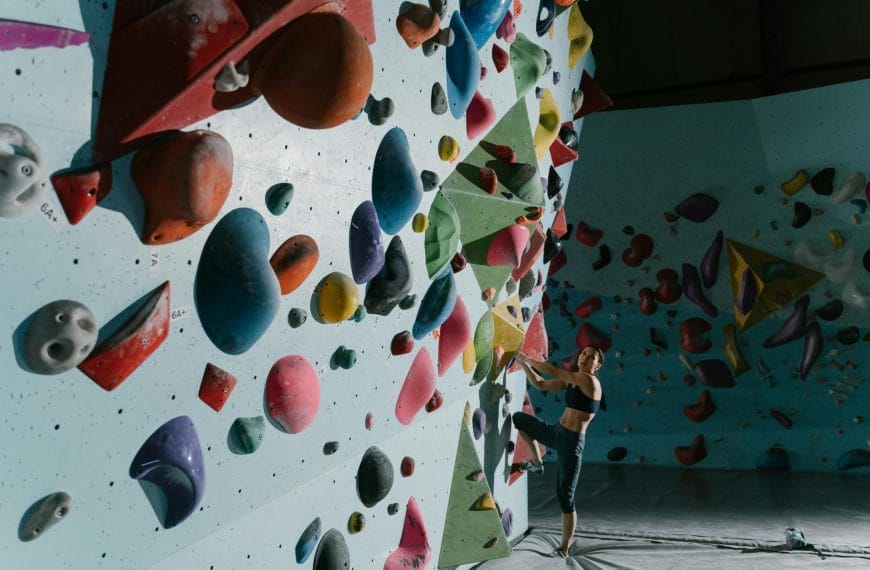 A colorful indoor rock climbing wall with a variety of differently shaped and sized climbing holds in various vibrant hues. A person is seen climbing on the wall.